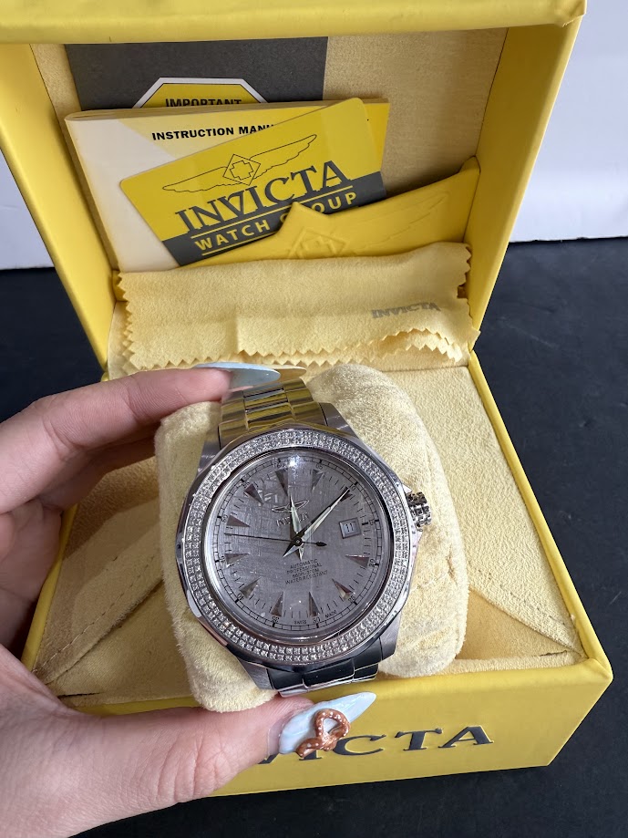 Silver Invicta wristwatch with a diamond-studded bezel displayed in a yellow presentation box, with manuals and a cleaning cloth nearby.