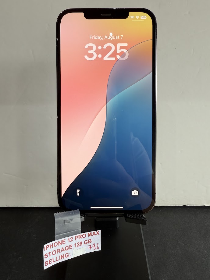 iPhone 12 Pro Max on a display stand in a store, price tag reads 'IPHONE 12 PRO MAX STORAGE 128 GB SELLING: -7%'.