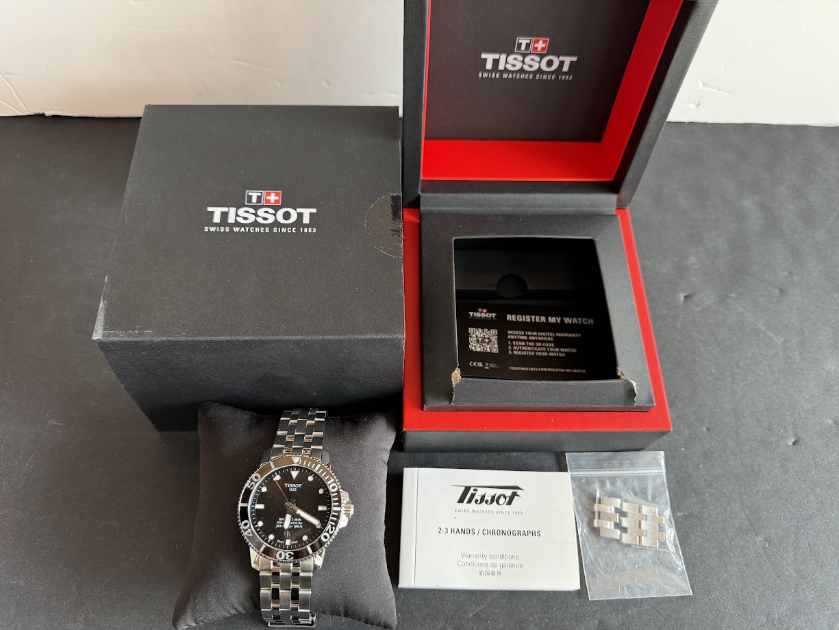 Tissot wristwatch with metal bracelet on a black pillow, beside its black box with white logo and red inner box contents displayed (manuals and extra links).